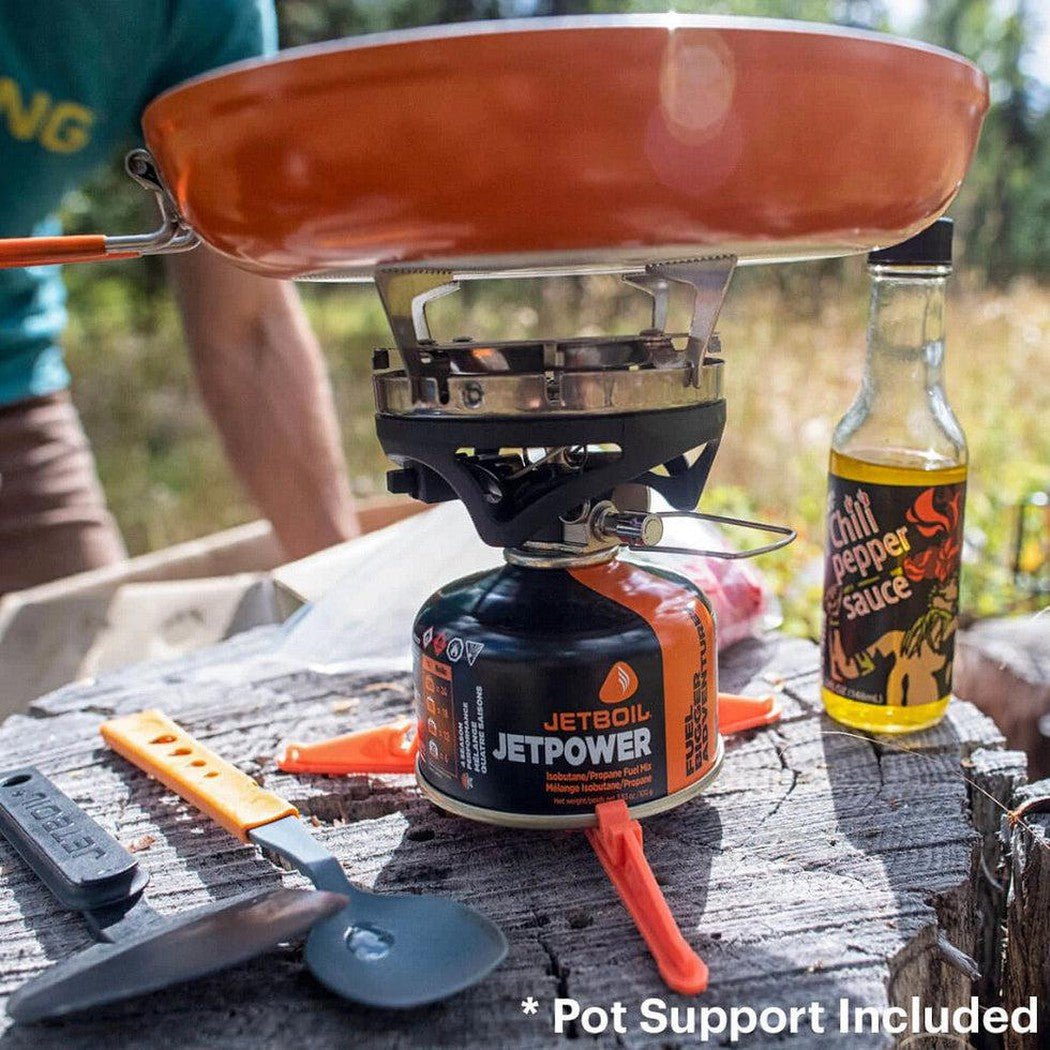 A top-down view of the JetBoil SUMO Cooking System on a log with a chili pepper sauce bottle nearby.