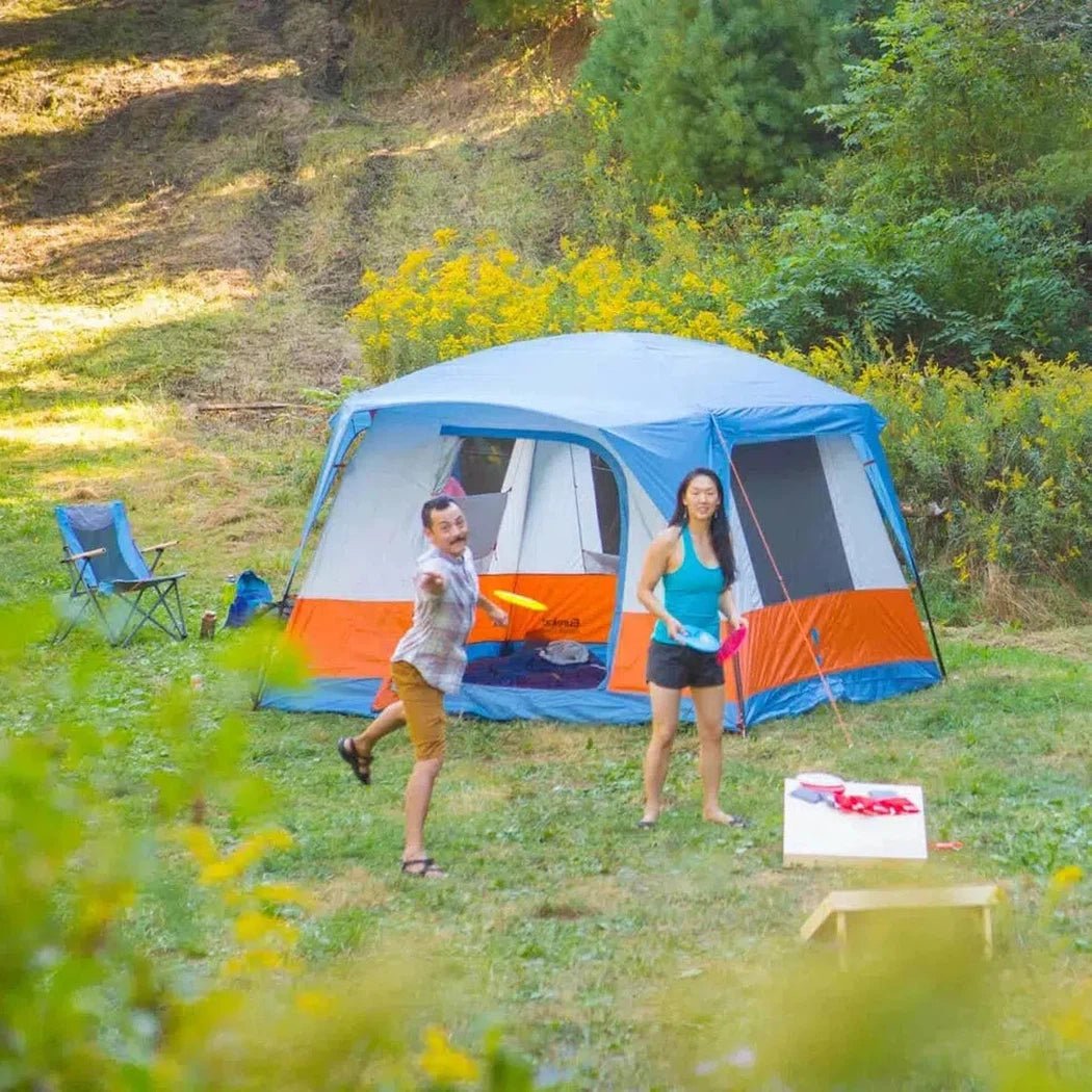 A man and woman play near a blue and orange Johnson Outdoors Eureka! Copper Canyon LX 4 tent in a grassy, wooded area.