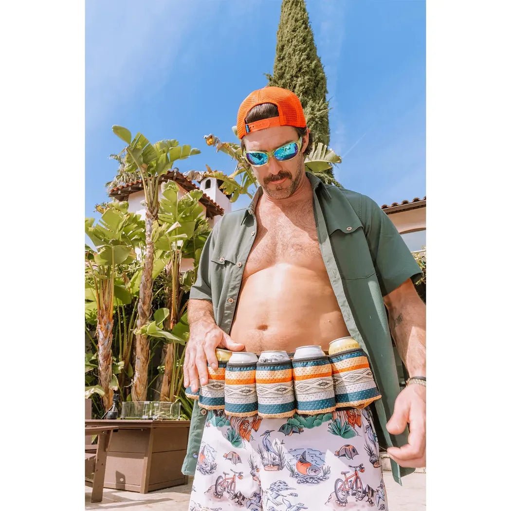 A man wearing the Kavu Beer Buddy in Park Lines stands outdoors with tropical plants and a cypress tree in the background.