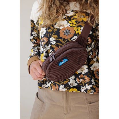 A model wears the Kavu Cord Spectator in Chocolate, a brown corduroy fanny pack with a blue logo, against a floral top and beige pants.