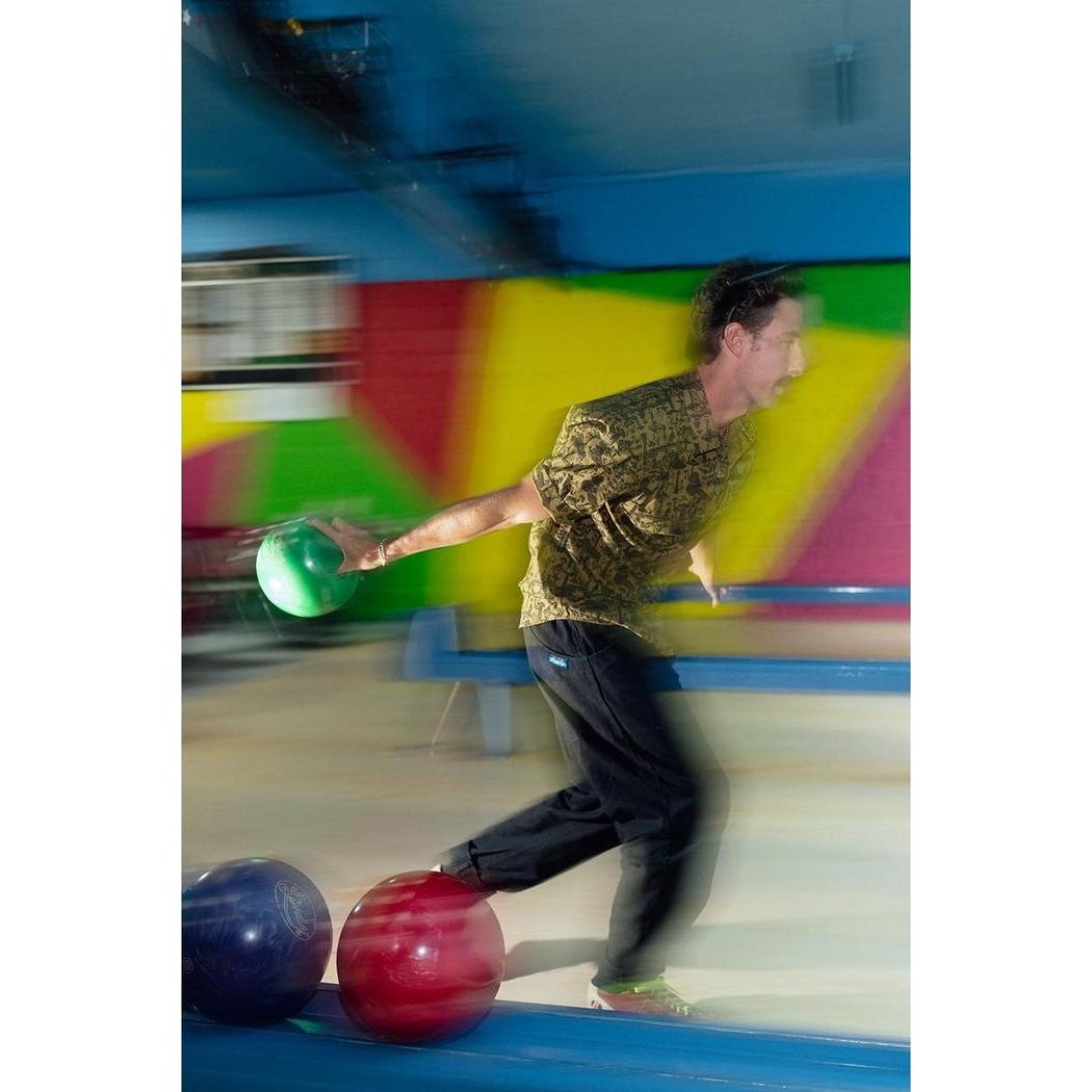 A man in a Kavu Men's Festaruski top bowls with a green ball at a colorful alley.