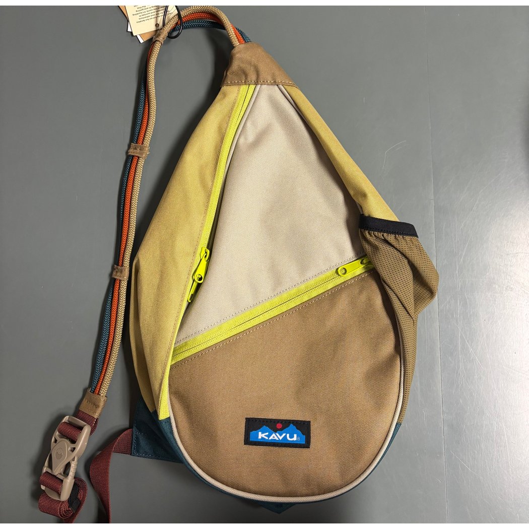 A front view of the Kavu Mini Paxton Pack in Forest Pass showing the yellow zipper and adjustable strap against a gray background.