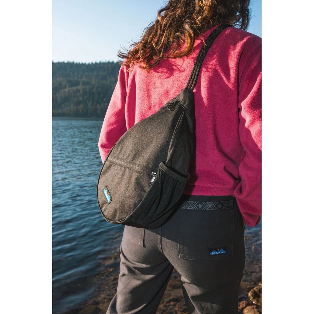A woman in a pink sweatshirt wears the Kavu Mini Paxton Pack in Forest Pass color, standing by a lake with trees in the background.