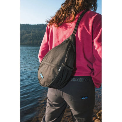 A woman in a pink sweatshirt wears the Kavu Mini Paxton Pack in Forest Pass color, standing by a lake with trees in the background.