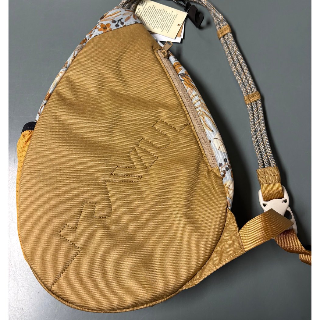 A top-down view of the Kavu Mini Paxton Pack in Forest Pass with a floral pattern on the back panel and a tan front pocket, against a gray background.