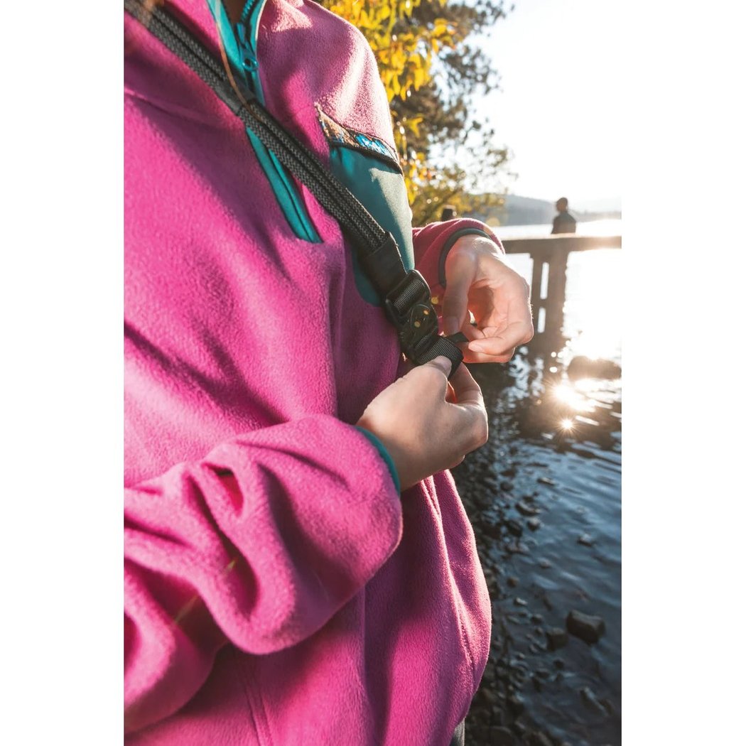 A person in a pink fleece adjusts the Kavu Mini Paxton Pack near a lake with a dock in the background.