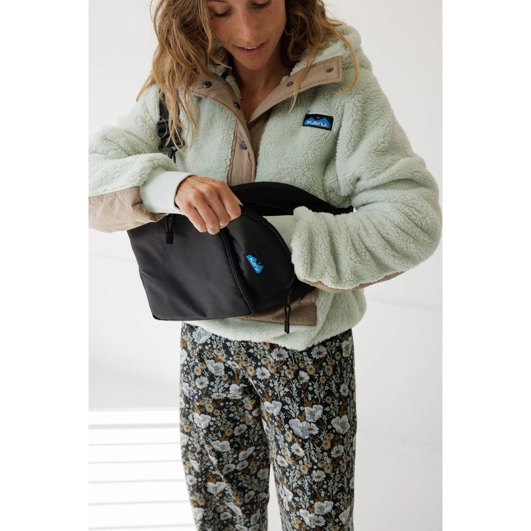 A woman wearing a Frosty Foliage jacket holds the Kavu Pender Pack against a white background.