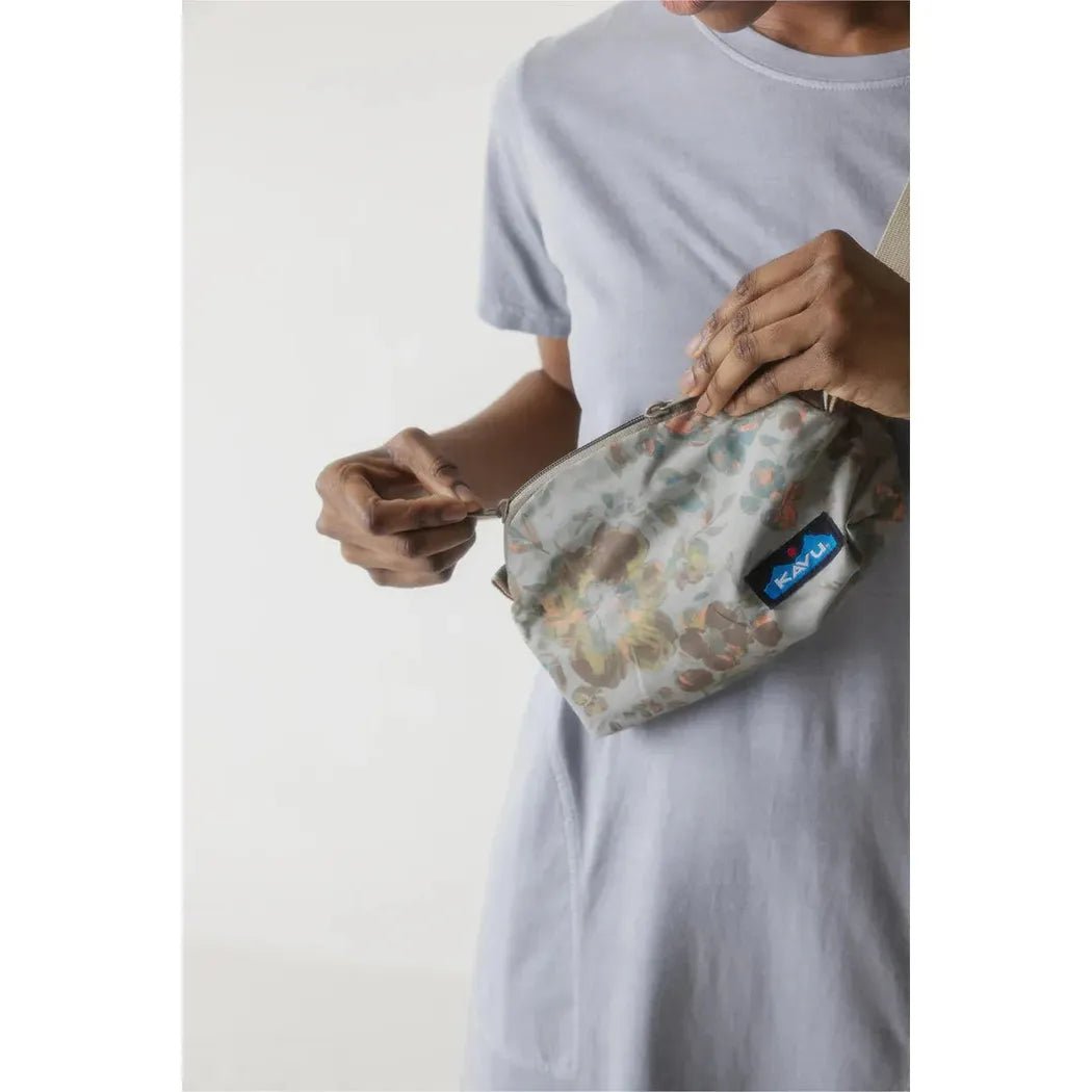 A person wearing a light gray t-shirt holds the Kavu Pint Size bag in Calm Palm color, showing the front with a blue tag, against a plain white background.