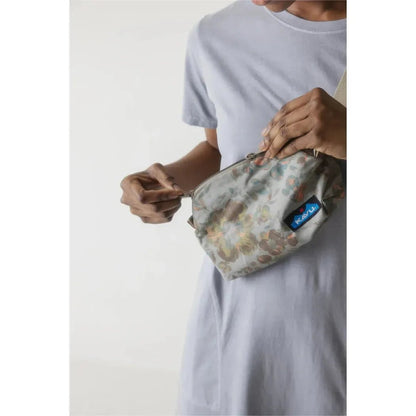 A person wearing a light gray t-shirt holds the Kavu Pint Size bag in Calm Palm color, showing the front with a blue tag, against a plain white background.