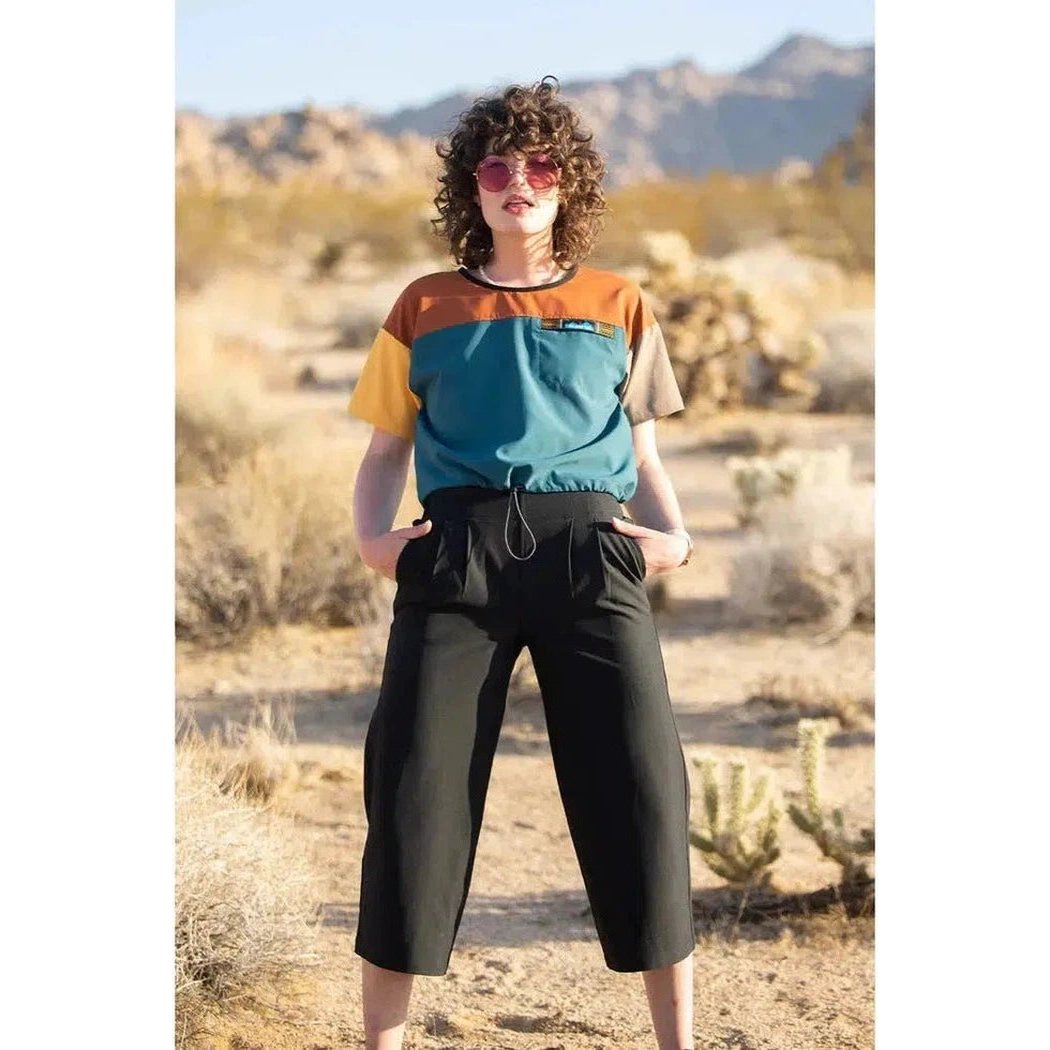 A woman wearing the Kavu Women's La Ticia Black cropped pants stands in a desert landscape with cacti and mountains behind her.