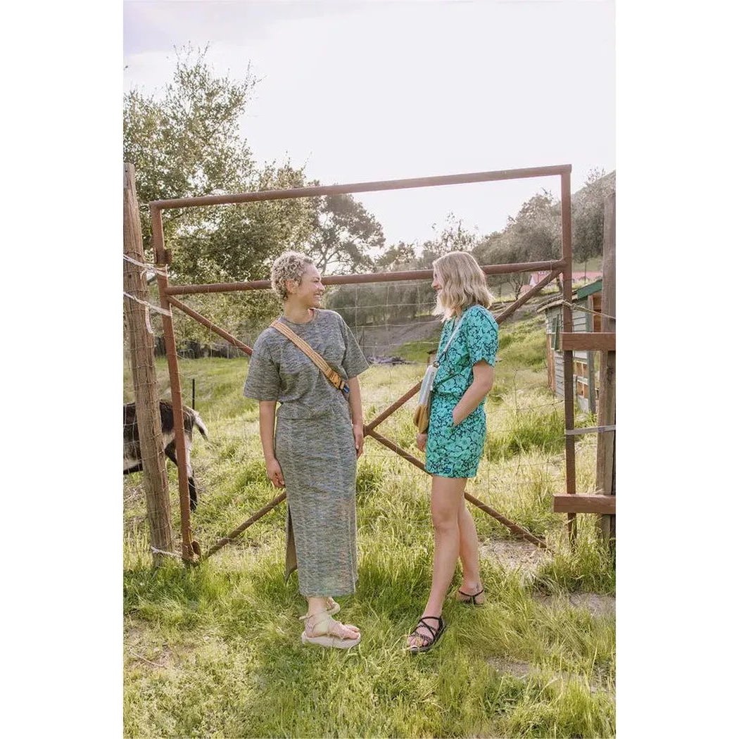 Two women wearing Kavu Women's Nosara Green Bloom bottoms chat near a rustic metal gate in a grassy outdoor setting.