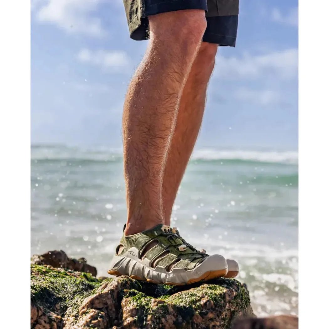A man stands on a mossy rock by the ocean wearing the Keen Men's Hyperport H2 in Martini Olive/Plaza Taupe.