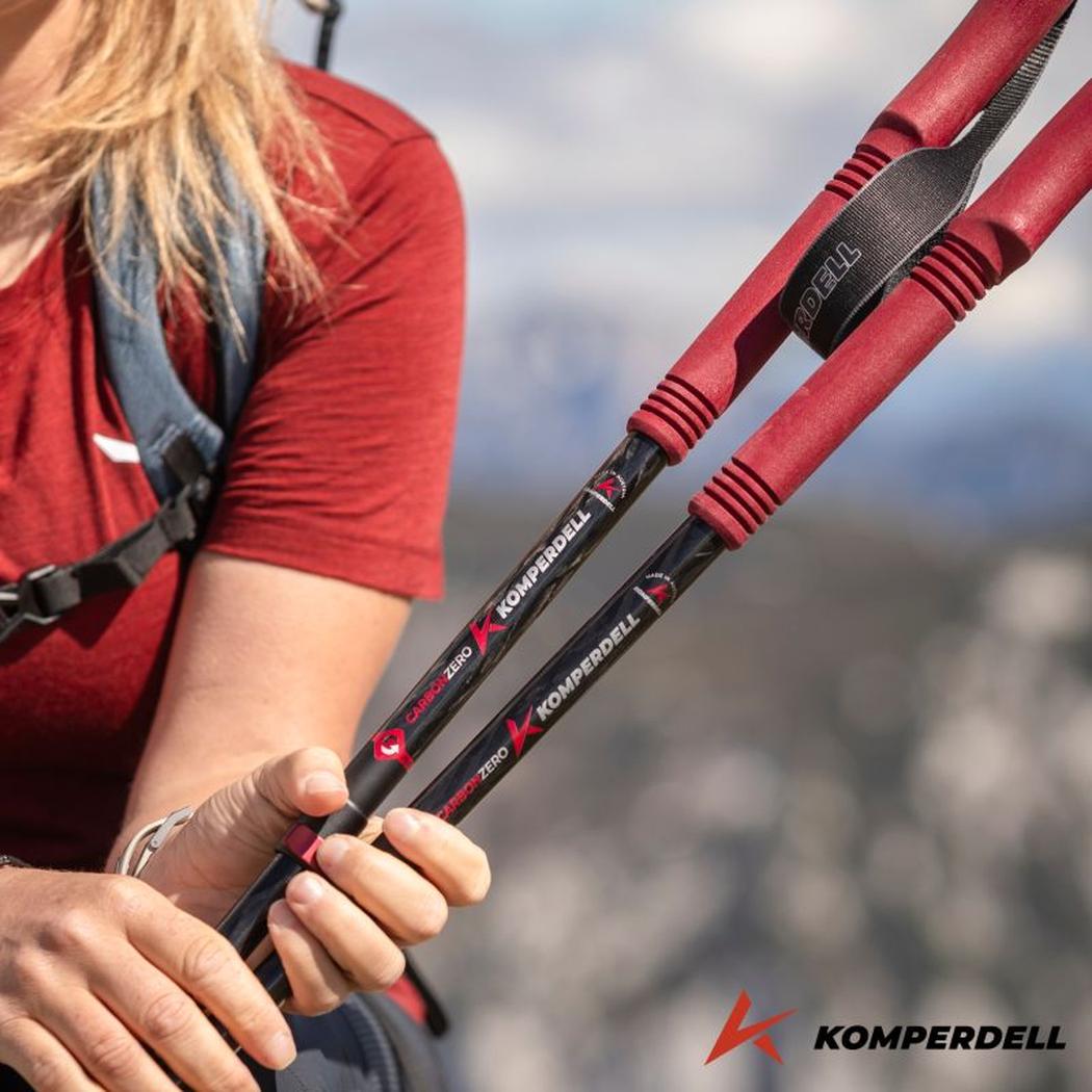 A woman holds Komperdell Carbon PRO ZERO Compact trekking poles with red grips against a mountain backdrop.