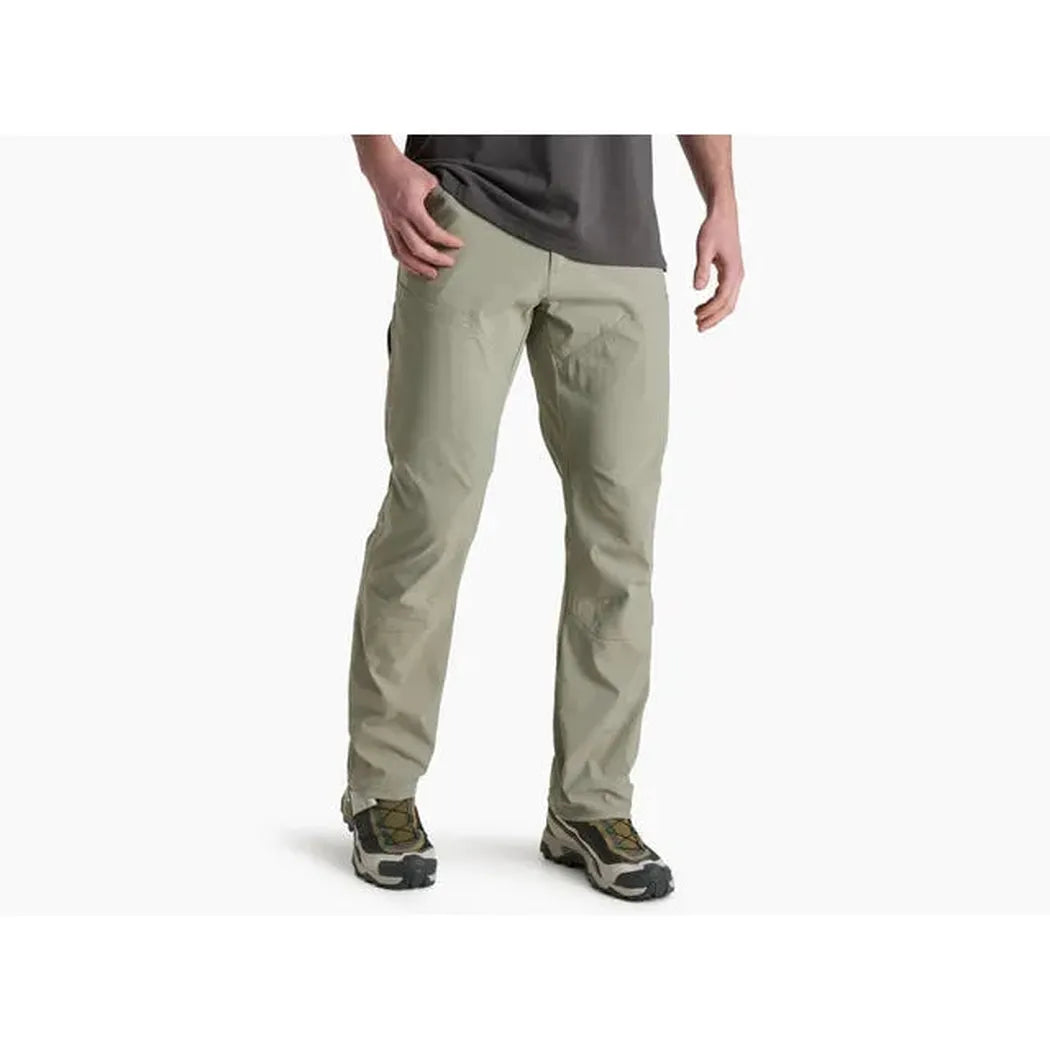 Side view of the Kuhl Men's Renegade Pant in Buckskin Khaki on a model wearing hiking shoes against a white background.