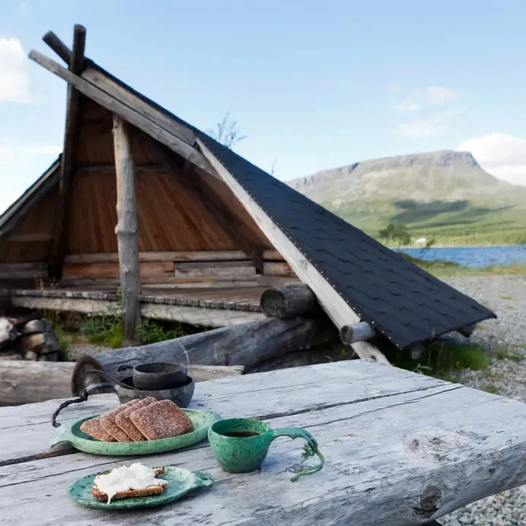 A rustic wooden table holds a black Kupilka 14 Small Plate with bread, a green mug, and a slice of toast, set against a log cabin and mountain backdrop.