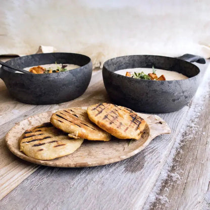 A close-up of two black Kupilka 14 Small Plates with soup and a wooden plate of grilled flatbread on a weathered wood table.
