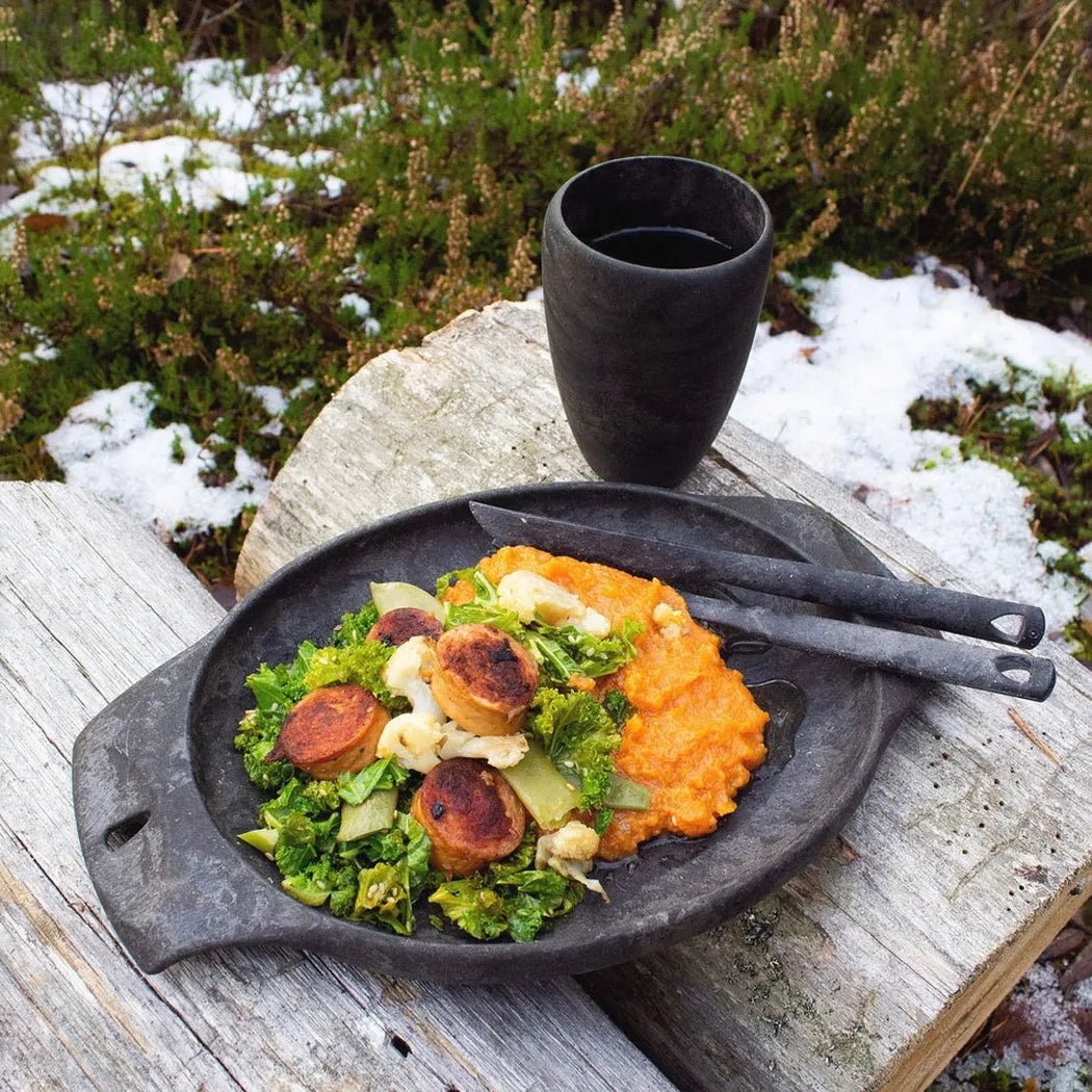 A black Kupilka 30 Tall Cup sits on a weathered wooden table with a meal of greens and sweet potato mash, surrounded by snow and greenery.