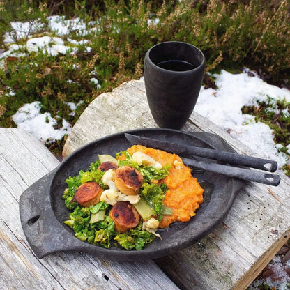 A black Kupilka 30 Tall Cup sits on a weathered wooden table with a meal of greens and sweet potato mash, surrounded by snow and greenery.