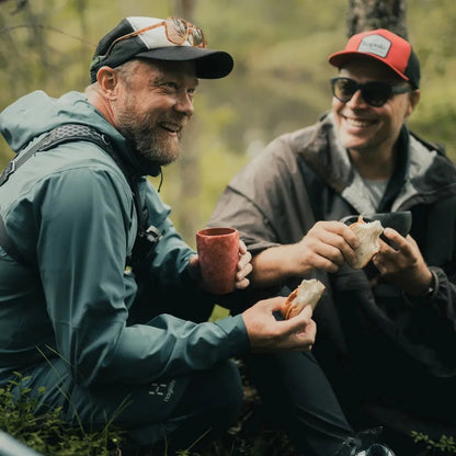 Two men in outdoor gear sit outdoors, one holding a red Kupilka 30 Tall Cup.
