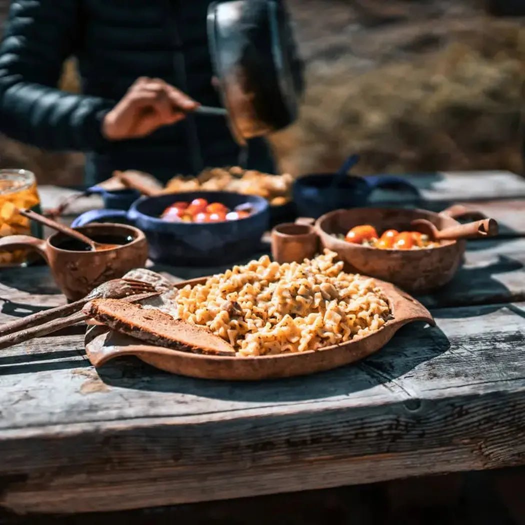 A person in a black jacket cooks over an open fire while a Kupilka 44 Large Plate holds mac and cheese on a weathered wooden table.