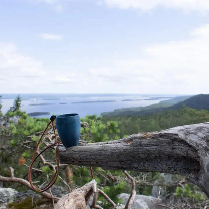 A black Kupilka 5 Shot Cup rests on a weathered tree branch overlooking a scenic lake and forest landscape.