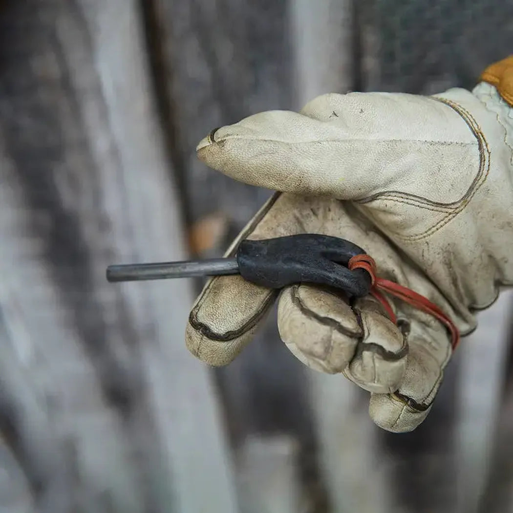 A gloved hand holds the Kupilka Firesteel 8 with a black handle and red cord, against a blurred wooden background.