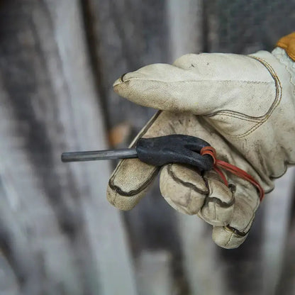 A gloved hand holds the Kupilka Firesteel 8 with a black handle and red cord, against a blurred wooden background.