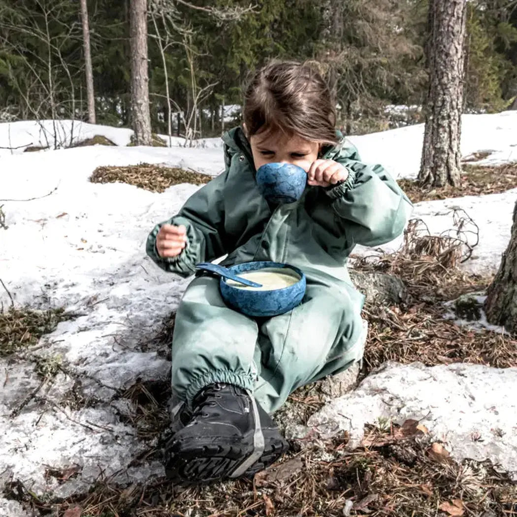 A young child in a green snowsuit sits on snowy ground with a blue bowl and the Kupilka Spork 165 Junior Utensil, surrounded by trees.