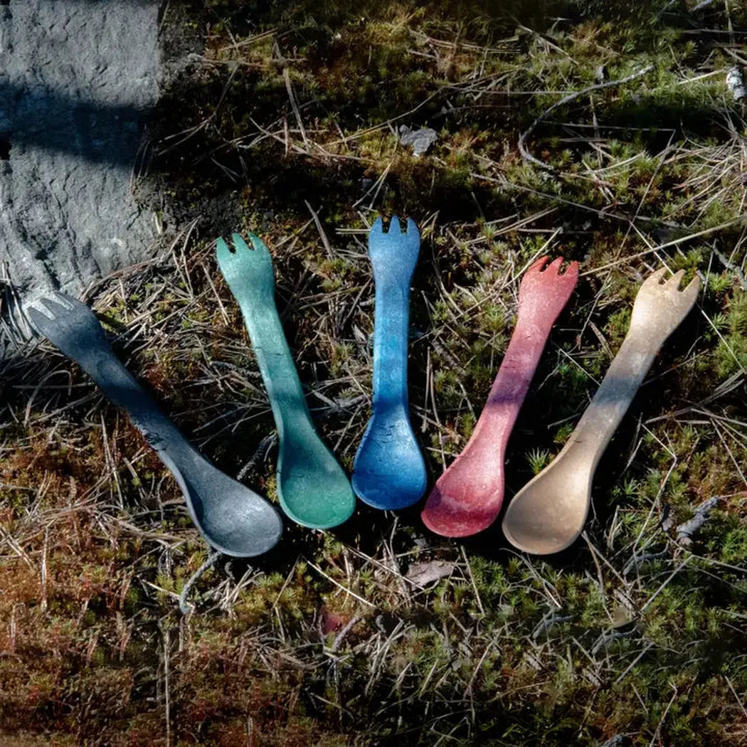 Five Kupilka Spork 165 Junior Utensils in Charcoal/Graphite, Teal, Blue, Pink, and Tan lie on moss and pine needles near a rock.