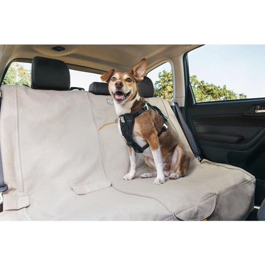 A dog wearing a harness sits on a black Kurgo Wander Bench Seat Cover in a car with a beige seat cover and trees outside.