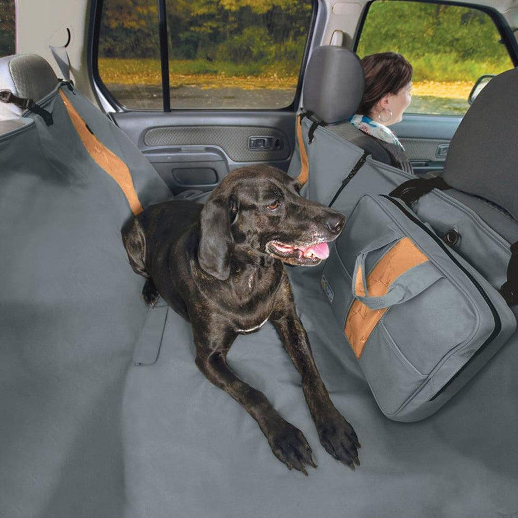 A black dog lies on a Charcoal Kurgo Wander Hammock in the back of a car, with a gray bag and a woman in the front seat visible.