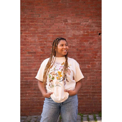 A woman wearing the Natural Liberty Graphics Mushrooms Short Sleeve T stands in front of a red brick wall.