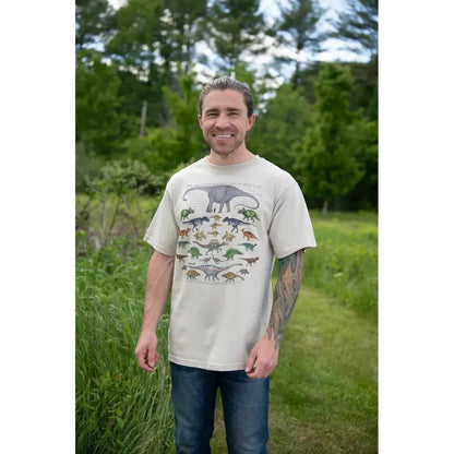 A man wearing the Stone Liberty Graphics One Dinosaur Is tee stands on a grassy path with trees in the background.
