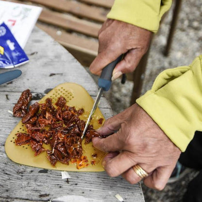 A person in a yellow jacket uses the Light My Fire Cutting Board BIO 2in1 to chop sun-dried tomatoes on a weathered outdoor table.