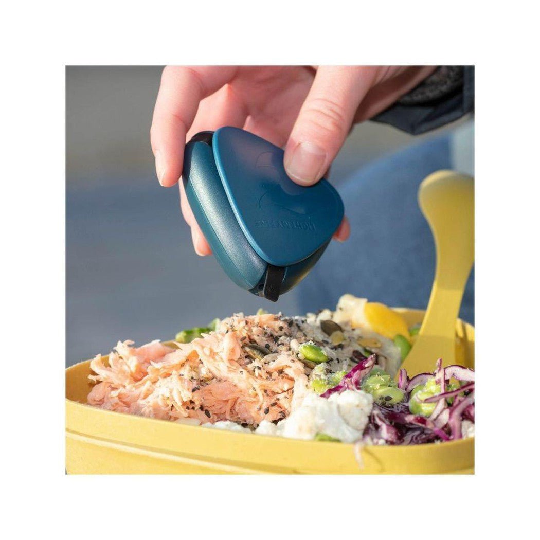 A hand uses the Dusty Pink Light My Fire Salt&Pepper plus BIO to season a colorful salad in a yellow container outdoors.