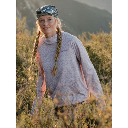A woman wearing the Marmot Women's AirExchange SolarShield Hoody in Dust Twirl stands among bushes with mountains in the background.