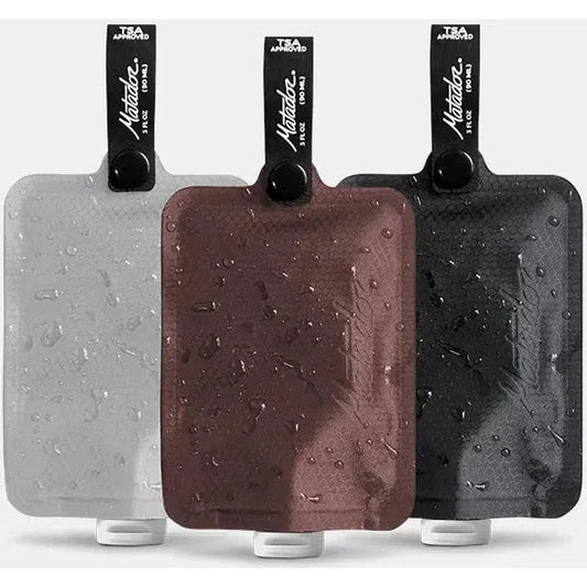 Three Matador FlatPak Toiletry Bottles in Charcoal/Garnet/Arctic with water droplets on their surfaces.