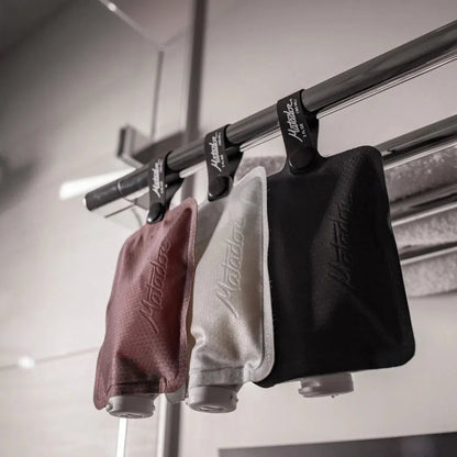Three Matador FlatPak Toiletry Bottles in Charcoal/Garnet/Arctic hang from a metal rod in a clean, modern space.