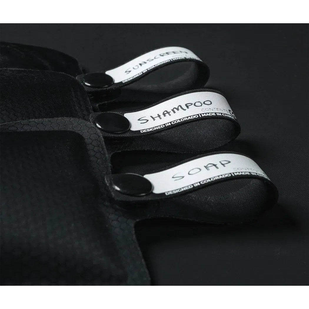 A close-up of the Matador FlatPak Toiletry Bottle (3-Pack) in Charcoal/Garnet/Arctic showing the shampoo, soap, and sunscreen straps against a dark background.