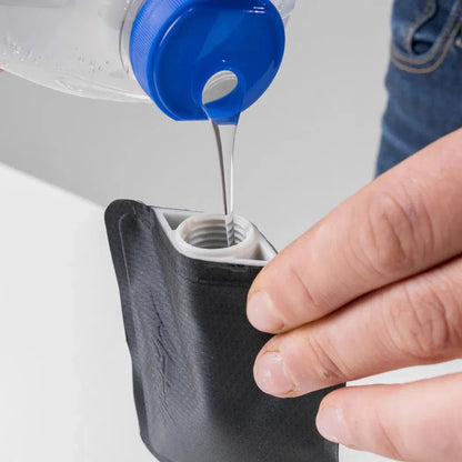 A hand pours liquid from a blue-capped bottle into a charcoal-colored Matador FlatPak Toiletry Bottle.