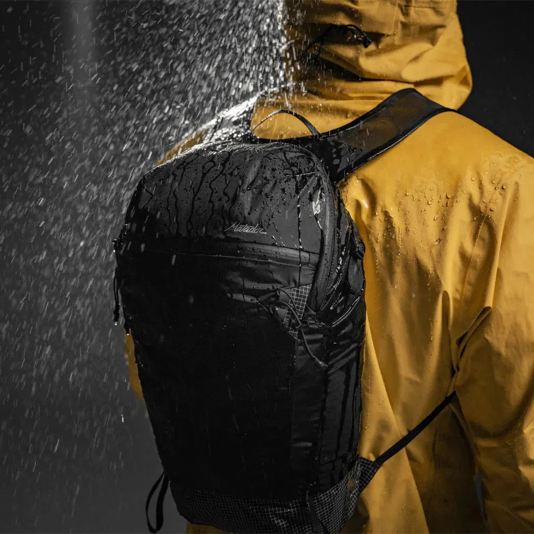 A person in a yellow jacket wears the black Matador Freefly 16 Packable Backpack as water sprays over it.