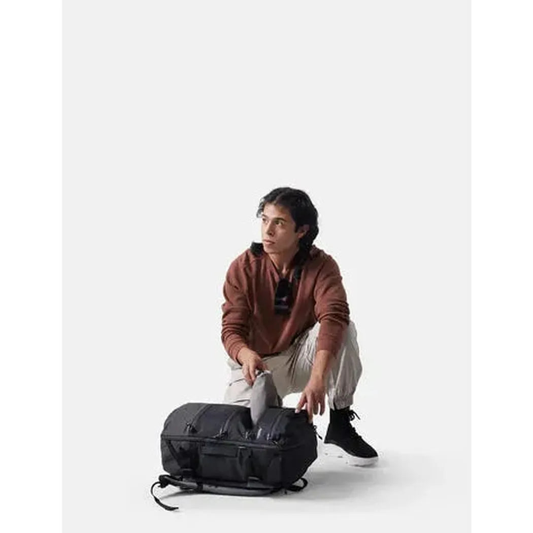 A model crouches to pack a black Matador SEG28 Backpack against a plain white background.
