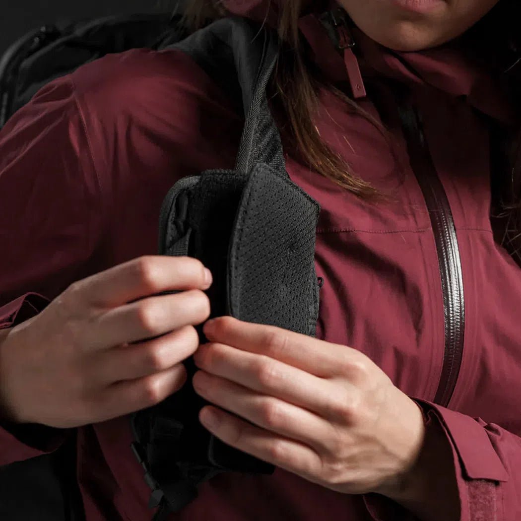 A close-up of a woman in a maroon jacket adjusting the black Matador Speed Stash backpack strap.