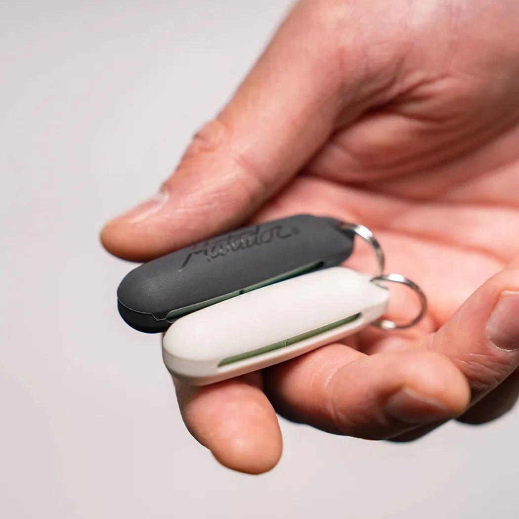 A hand holds the Matador Travel Earplugs Kit in charcoal and white, showing the compact design and metal keyring attachment.