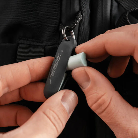 A person holding the Matador Travel Earplugs Kit, showing the charcoal-colored case and light green earplugs against a black backpack.
