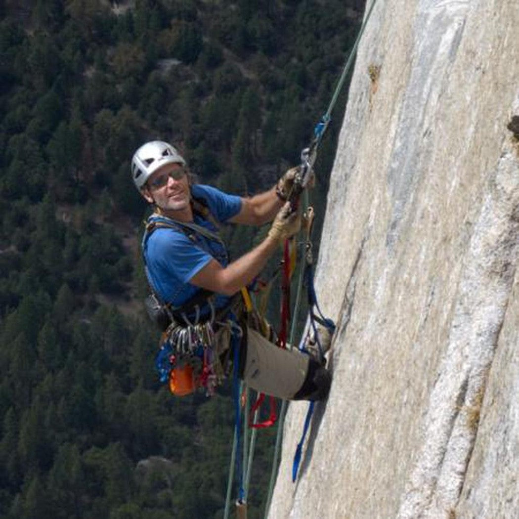A climber wearing Metolius Climbing Glove - 3/4 Finger gloves scales a rock face with trees in the background.