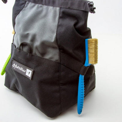 A blue Metolius Razorback Boar's Hair Brush hangs from the side of a black and gray Metolius bag against a white background.