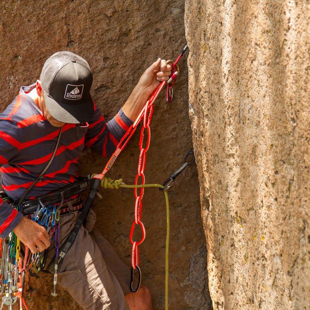 A climber in a striped shirt and charcoal cap uses the Metolius Ultimate Daisy Chain in Blue/Green to secure themselves to a rock face.