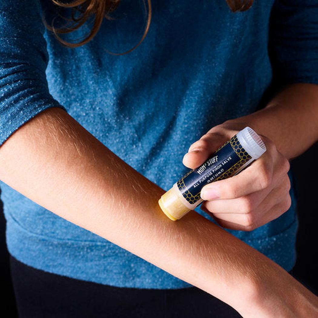 A woman in a blue sweater applies Moms Stuff Salve All Purpose Pinon Salve to her arm in a close-up shot.
