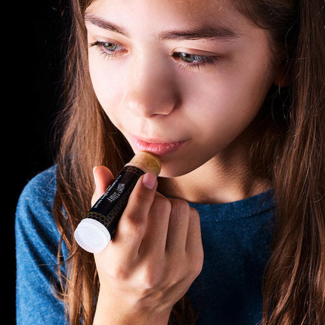 A young girl in a blue shirt holds the Moms Stuff Salve All Purpose Pinon Salve - Large Tube close to her lips against a dark background.